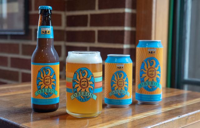 An assortment of Bell's Oberon Ale products, including a glass bottle, a filled pint glass, and two sizes of cans, featuring the brand's signature orange sun logo on a blue and yellow background.