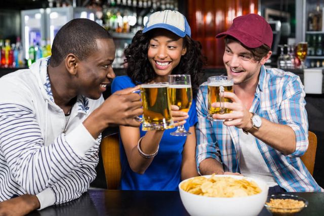 A smiling image of three friends (two men and a woman) clinking their beer mugs and having bowls of snacks on the table at a bar.