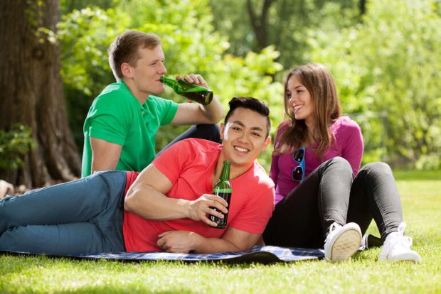 Three college students sitting on grass on campus, chatting and drinking beer together outdoors.