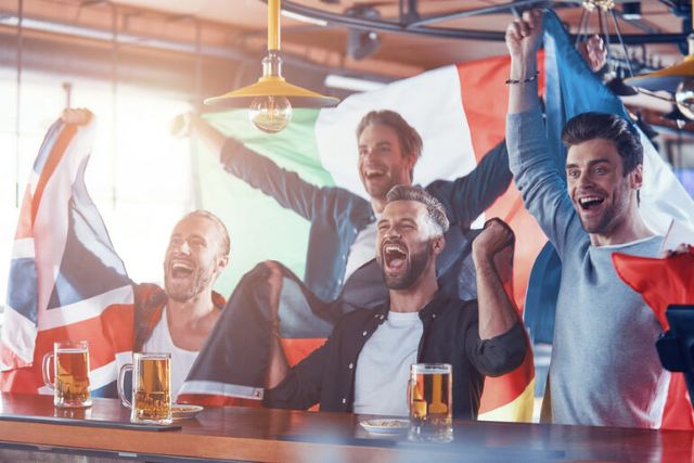 Cheering fans at a bar waving international flags with beers on the counter.