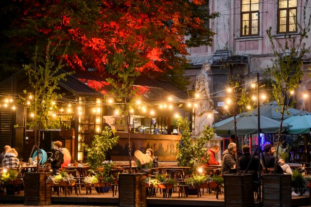 An outdoor urban beer garden at night with people seated at tables under warm string lights, featuring a central bar structure, potted greenery, and a large tree illuminated with red light.