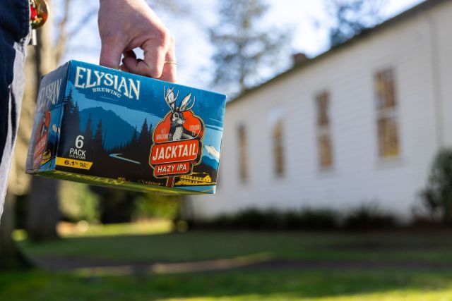 A hand holding a six-pack of Elysian Brewing Jacktail Hazy IPA in front of a white building.
