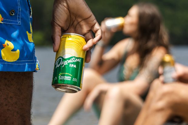An outdoor, sunlit scene where a close-up, hand-held yellow and green can of Daycation IPA is the focus. In the blurred background, people in swimwear lounge and drink by a body of water.