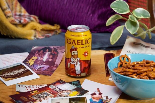 A yellow can of Highland Brewing Gaelic Amber Ale sits on a wooden table alongside a blue bowl of pretzels, scattered printed photographs, and a green potted plant.
