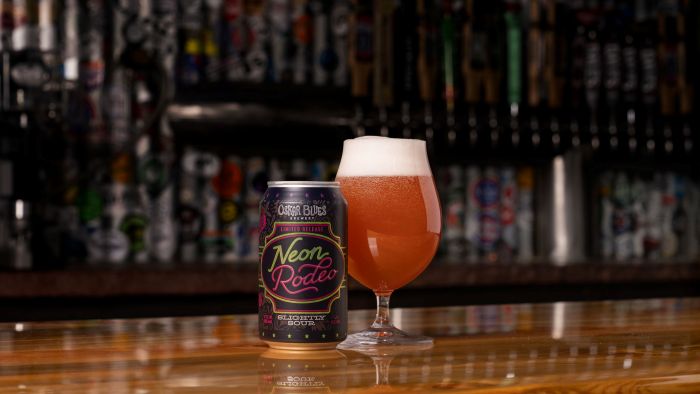 A can of Oskar Blues Neon Rodeo beer sits on a wooden bar next to a filled, foamy glass..