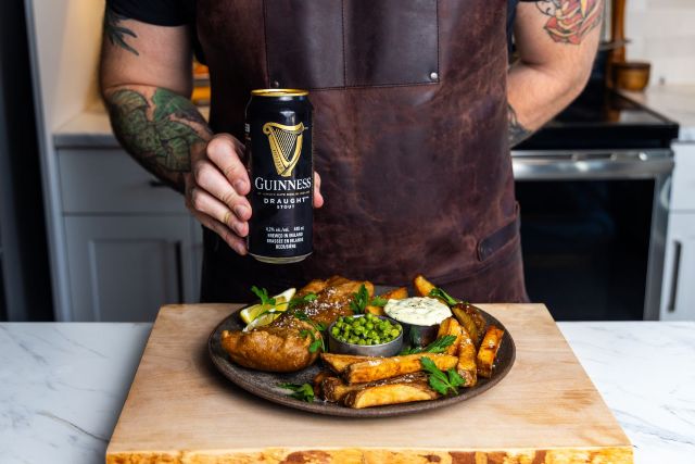 A person in a leather apron holding a can of Guinness Draught Stout behind a plated meal of fish and chips with peas and tartar sauce.