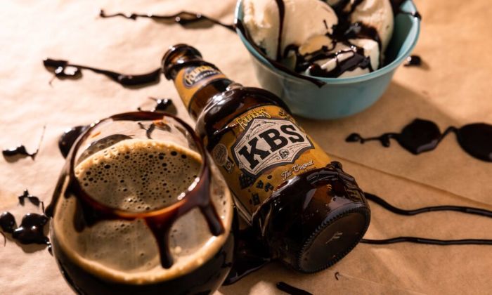 A bottle of Founders KBS bourbon barrel-aged stout rests on its side next to a glass of the dark beer and a bowl of vanilla ice cream topped with hot fudge.
