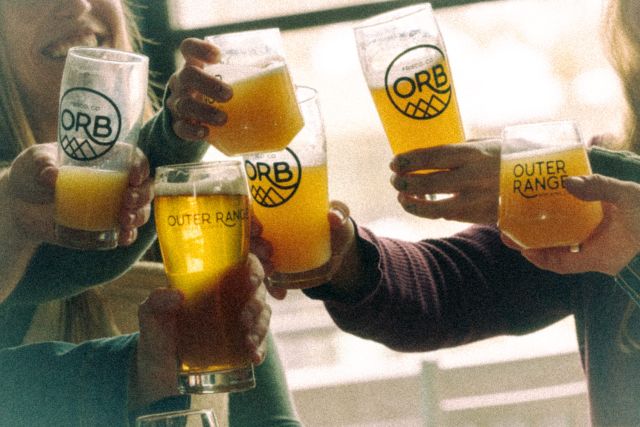 A group of friends toasting with glasses of beer, some bearing the "Outer Range Brewing" logo and "ORB" emblem.