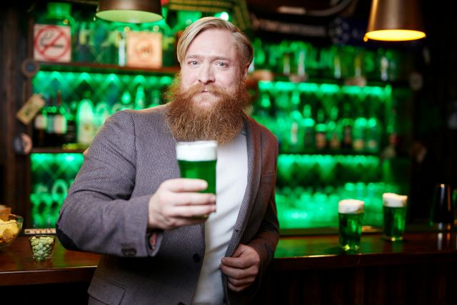 A bearded man in a blazer holds up a glass of green beer in a pub decorated with green lights for St. Patrick's Day.