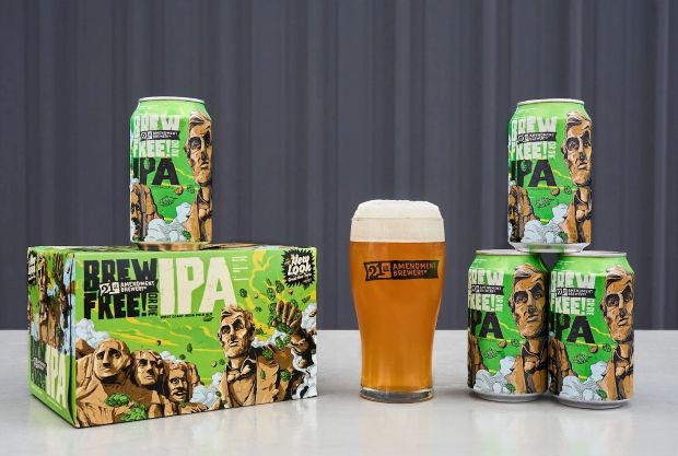 Several cans and a cardboard box of 21st Amendment Brewery's "Brew Free! or Die" IPA are arranged on a white surface next to a full, frothy glass of the beer. The bright green packaging features stylized illustrations of Mount Rushmore and Abraham Lincoln.