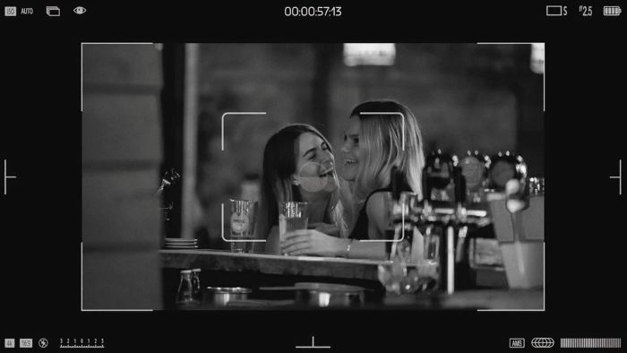 A black-and-white, candid shot framed like a camera viewfinder shows two women laughing together at a bar. 