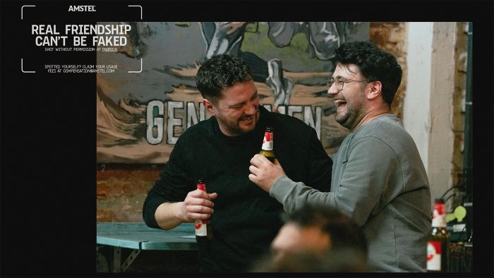 Two men share a laugh over beers in this candid photograph. One man, in a black sweater, holds a bottle of Amstel beer while smiling down at it. His companion, wearing a grey sweatshirt and glasses, laughs heartily with his head tilted back. A sign with the word "GENTLEMEN" is partially visible in the background, along with a mural. The image is framed with an Amstel logo and the text "REAL FRIENDSHIP CAN'T BE FAKED."