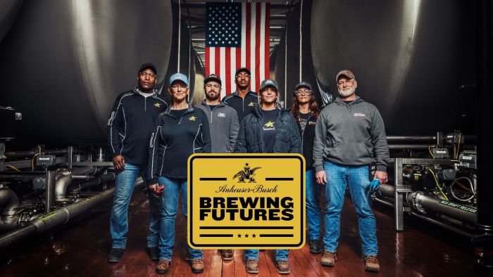 Seven employees stand in a brewery in front of a large American flag and brewing tanks, with a prominent yellow sign in the foreground reading "Anheuser-Busch BREWING FUTURES.
