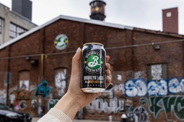 A hand holds a can of Brooklyn Lager in front of the brick brewery .