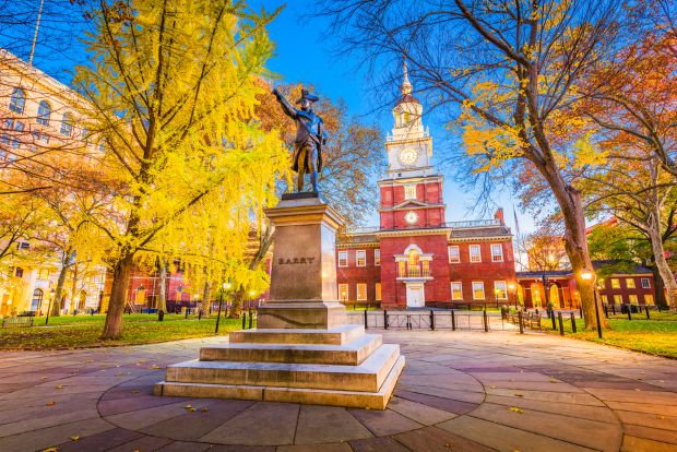 A statue of Commodore John Barry stands in the foreground, with the historic, red-brick Independence Hall and its clock tower under a blue twilight sky in Philadelphia.