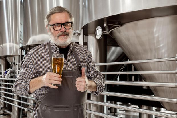 Brewer holding craft beer in a brewery with tanks.