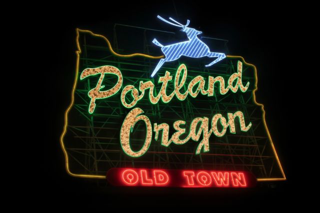 A neon sign at night in the shape of Oregon, featuring the words "Portland Oregon" in glowing script, "Old Town" in a red banner below, and a leaping white stag on top.