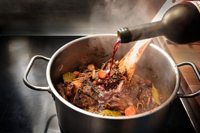 Bottle of red wine being poured into a steaming pot of beef stew with vegetables.