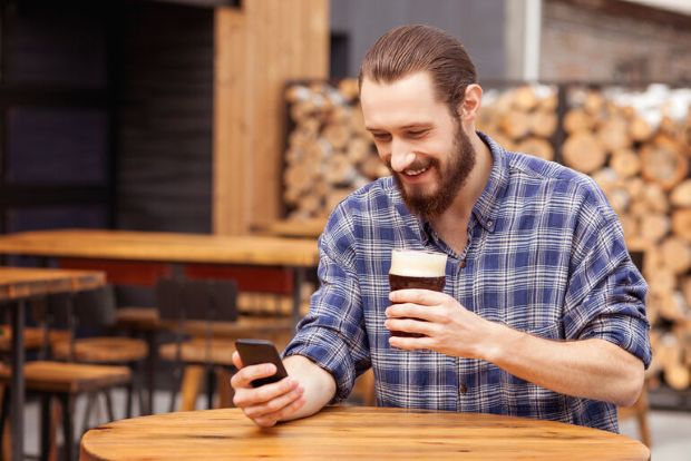 Man holding a craft beer and checking his phone at a bar, showing digital engagement in modern beer culture.