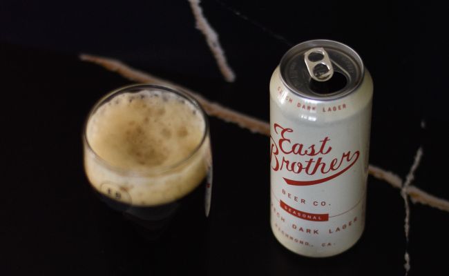 An open can of East Brother Beer Co. Czech Dark Lager next to a glass of dark beer with a thick head of foam.
