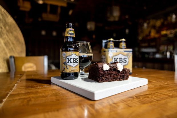 A bottle and glass of Founders KBS imperial stout sit on a wooden table next to a slice of chocolate cake with marshmallow filling. A four-pack of the same beer is visible in the background.