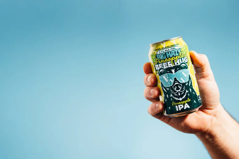 A person's hand holding a can of Goose Island "Big Hazy Pineapple Beer Hug" IPA against a light blue background.