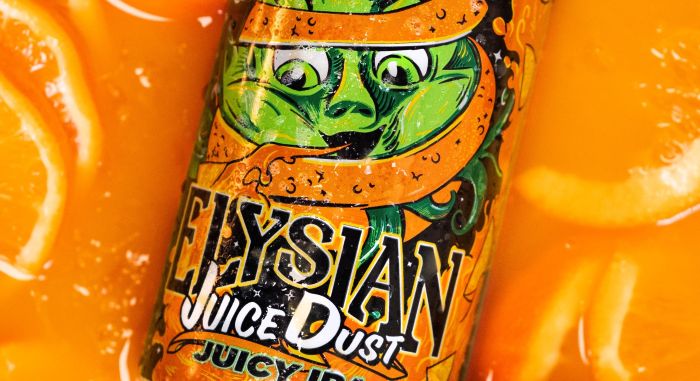 A close-up, angled view of an Elysian Juice Dust Juicy IPA can, partially submerged in bright orange juice with floating orange slices. The label features a cartoonish, green, wide-eyed character with an orange peel wrapped around its head.