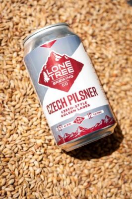 A can of Lone Tree Brewing Co. Czech Pilsner rests on a dense bed of malted barley grains. The can features a white, silver, and red label with mountain graphics.