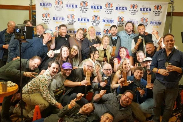 A large, diverse group of smiling people poses for a celebratory group photo in front of a white "BEER" and "Yakima Chief Hops" step-and-repeat banner. Many individuals are holding wooden awards, while others cheer and gesture toward the camera. The atmosphere is lively and casual, with several people kneeling or sitting in the front row of the crowded gathering.