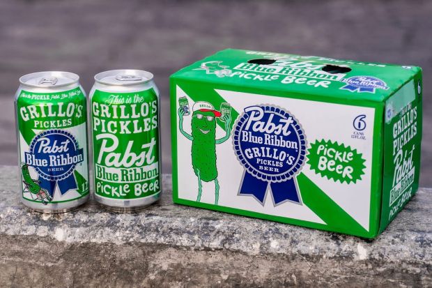 A 6-pack and two individual cans of Pabst Blue Ribbon x Grillo's Pickles "Pickle Beer" sit on a stone ledge. The packaging is bright green and white, featuring a cartoon pickle mascot wearing sunglasses and a hat
