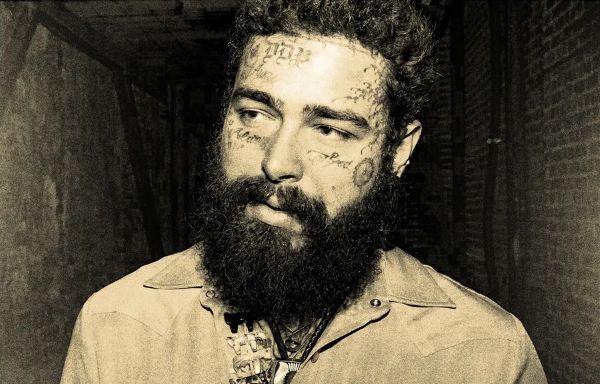 A sepia-toned, medium-up-close portrait of Post Malone, featuring his facial tattoos and a full dark beard, while he gazes off-camera.
