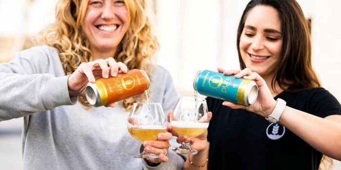 Two women smiling and pouring Pure Project beer from colorful cans into glass goblets.