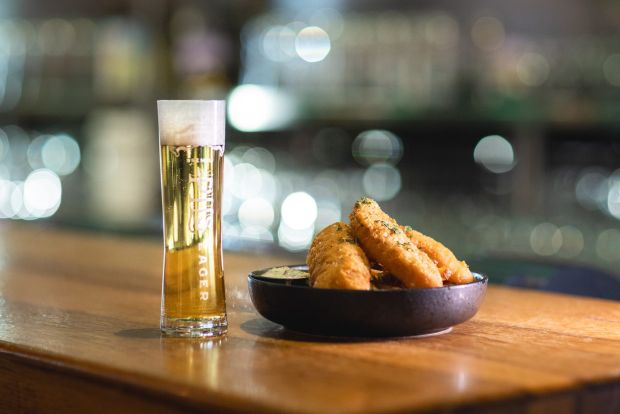 A tall glass of Sierra Nevad Premium Pils with a frothy head sits next to a bowl of crispy, golden-brown fried fish on a wooden bar top.