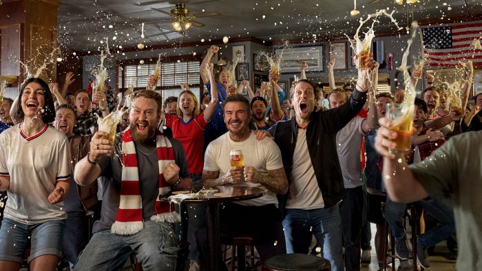 A crowded bar of cheering soccer fans, including David Beckham, celebrating with raised beer glasses amidst splashing foam and an American flag.