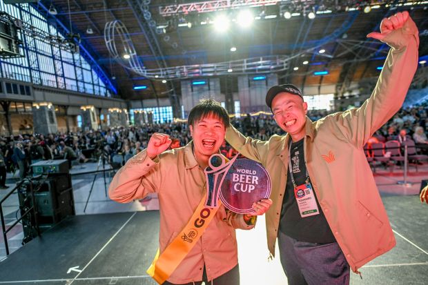 Two men smile and cheer while holding a World Beer Cup award on a stage in a large, crowded hall.