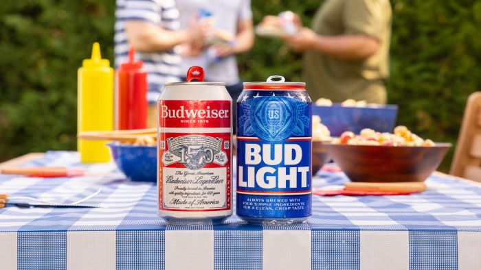 A can of Budweiser and a can of Bud Light sit side by side on a blue and white checkered picnic table at an outdoor gathering.