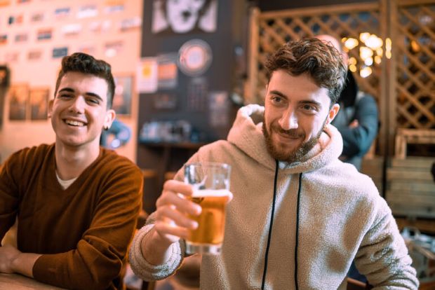 Two young men at a bar, one raising a glass of beer while smiling, capturing a casual social moment amid shifting Gen Z drinking habits.