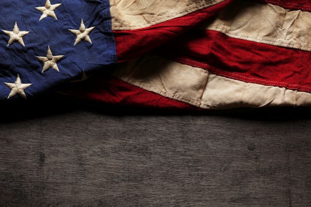 An American flag with an aged texture is draped horizontally across the top half of a dark wooden surface, leaving the bottom half open.