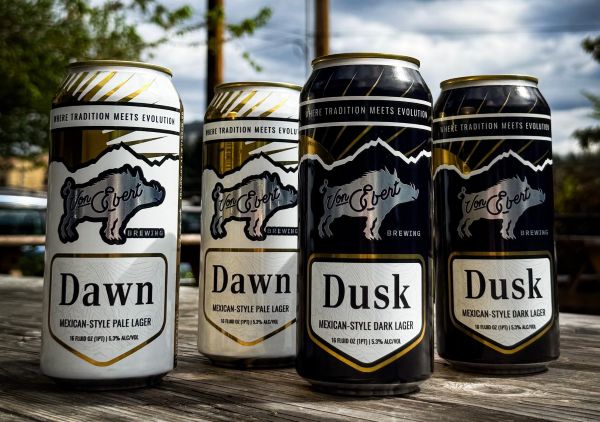 Four cans of Von Ebert Brewing's Dawn and Dusk Mexican-style lagers stand on a wooden surface outdoors. The two white cans on the left are Dawn Pale Lager, and the two black cans on the right are Dusk Dark Lager.
