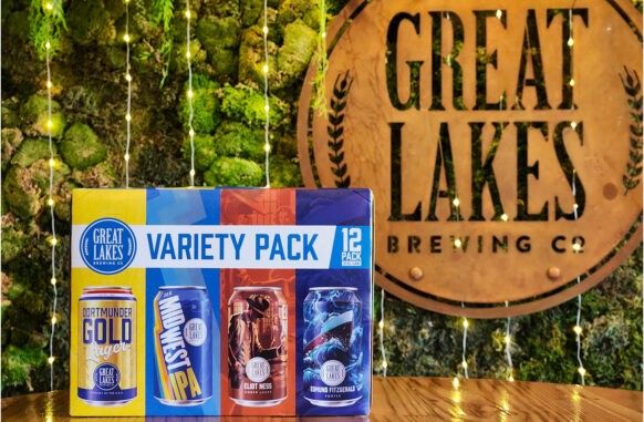 A 12-pack box of Great Lakes Brewing Co. Variety Pack sits on a wooden table next to a circular wooden brewery sign, set against a green mossy background with hanging fairy lights.