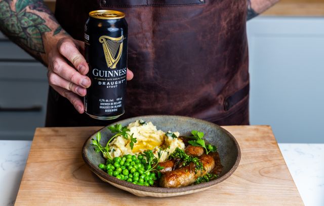 A person wearing a brown leather apron holds a tall, black can of Guinness Draught Stout in their right hand, positioning it above a rustic ceramic bowl filled with a classic meal of bangers and mash.