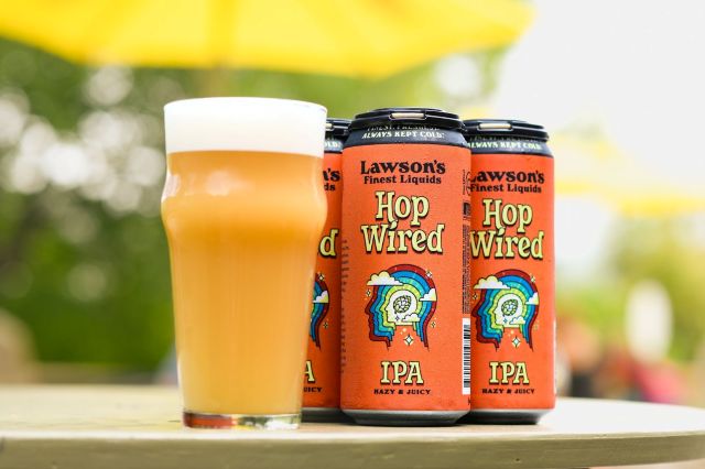 A glass of hazy, orange-colored beer with a white head stands in front of three orange cans of Lawson's Finest Liquids Hop Wired IPA.