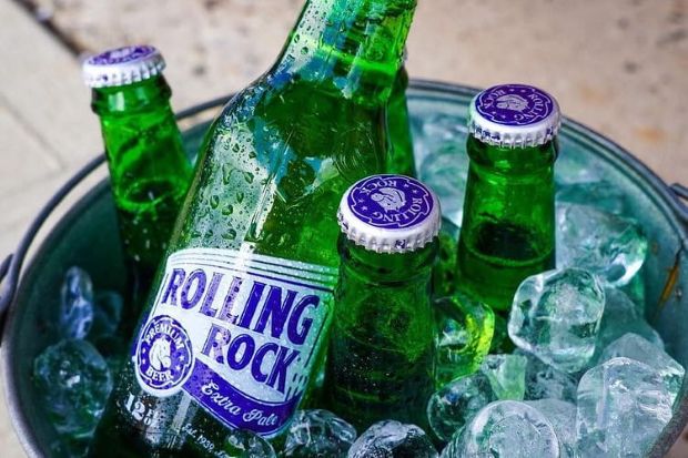 Several green glass bottles of Rolling Rock beer resting in a metal bucket filled with ice cubes.