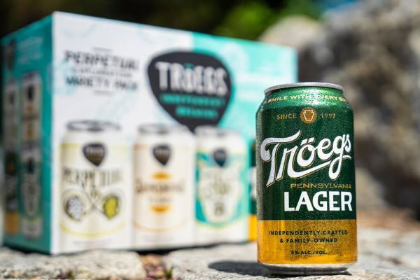 A green and gold can of Tröegs Pennsylvania Lager stands in sharp focus outdoors, with a blurry variety pack box of Tröegs beer in the background.