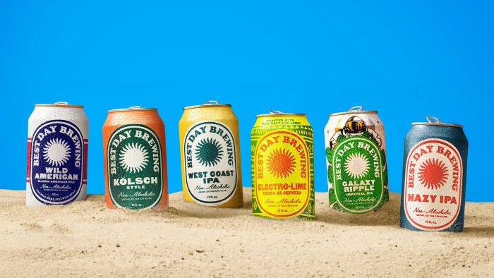 Six non-alcoholic craft beer cans by Best Day Brewing lined up on sand against a bright blue background.