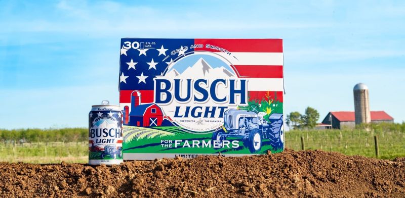 A 12-ounce can and a 30-pack box of Busch Light "For the Farmers" limited edition beer, featuring an American flag, tractor, and barn design, sitting outdoors on a mound of dirt with a real farm and silo blurred in the background.