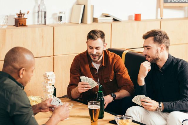 Friends playing cards with cash and beer on the table, showing how a poor betting site choice can spoil a relaxed game night.
