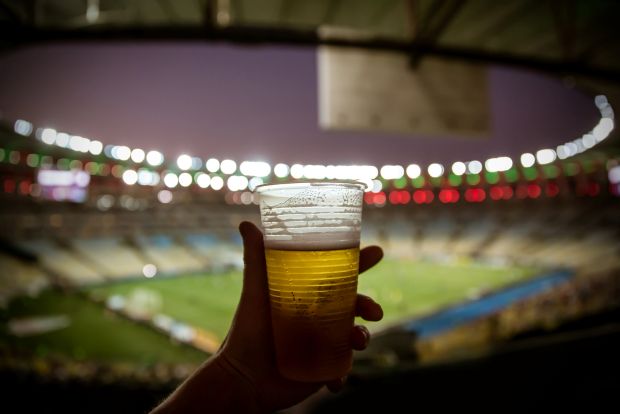 A hand holds a plastic cup of beer against the blurred background of a brightly lit soccer stadium at night.