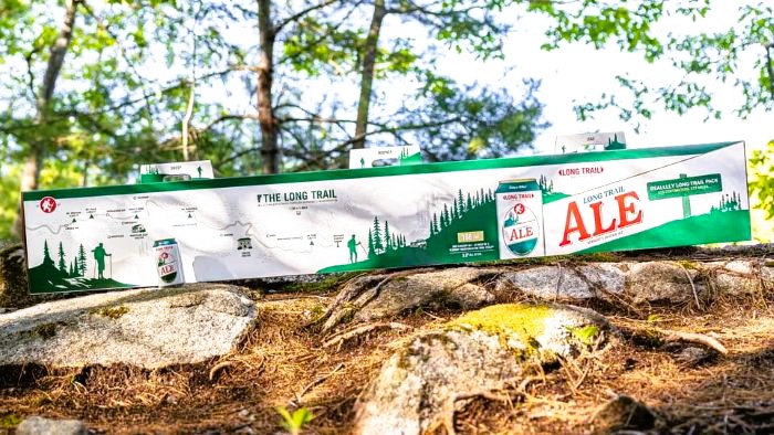 A long, green and white cardboard multi-pack box of Long Trail Ale beer sitting on mossy rocks outdoors.