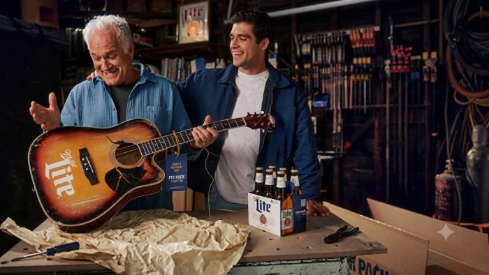 Father and grown-up son smiling together in a garage workshop, holding a broken acoustic guitar with a Miller Lite logo on it next to a six-pack of beer.